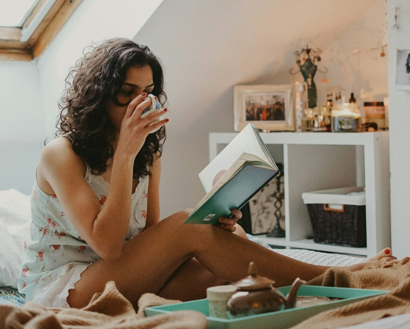 Woman in sleep clothes sitting on the bed sipping a cup of tea while reading a book
