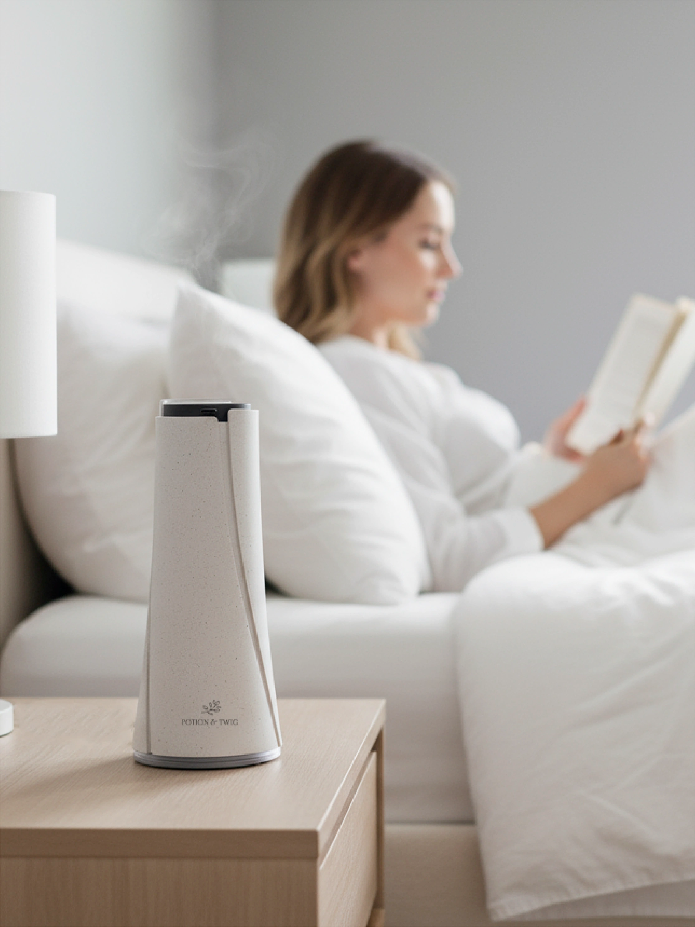 Woman reading a book on a bed with a white nightstand lamp and white Lumen wireless scent diffuser.