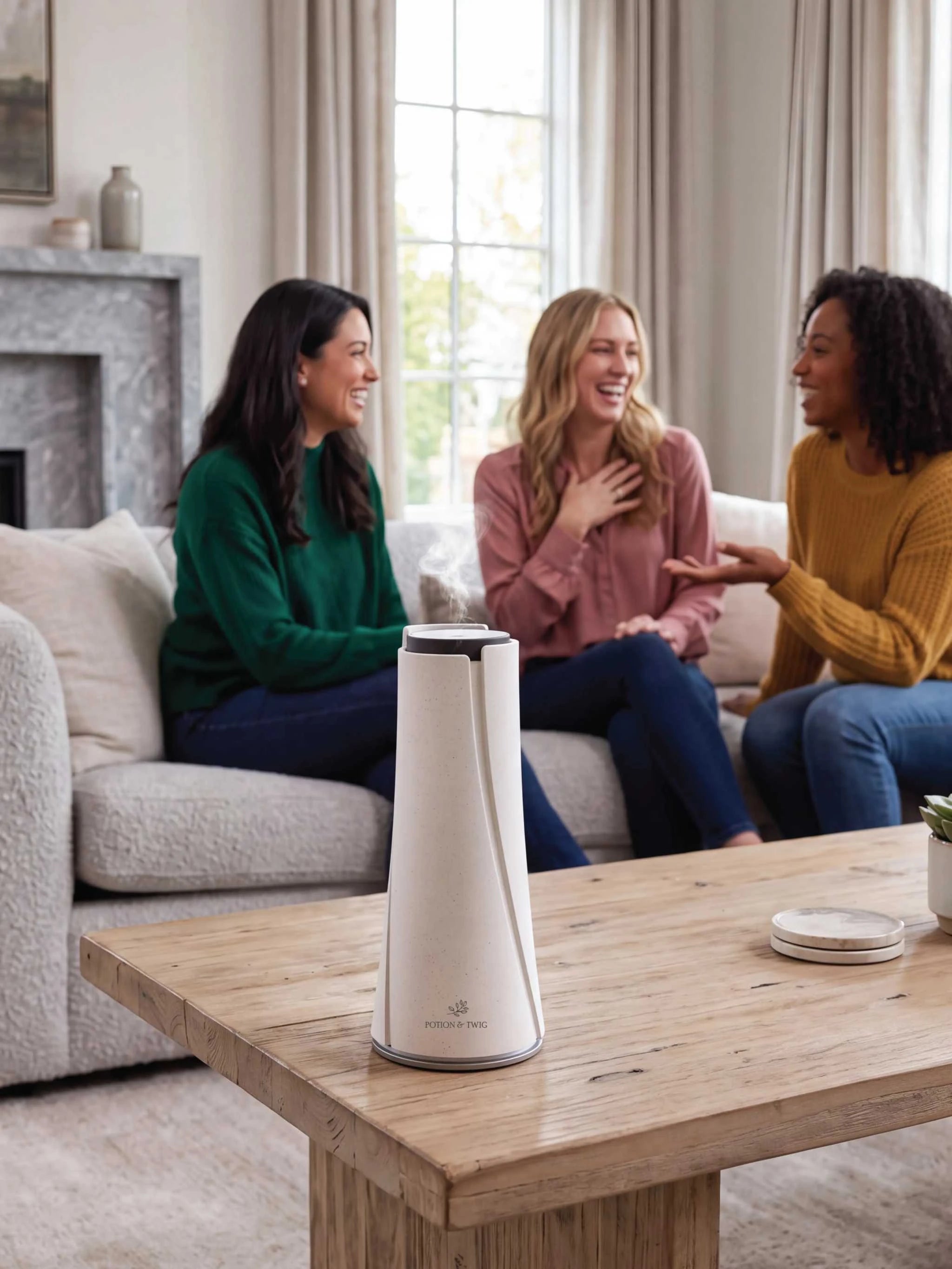 A white and silver countertop diffuser sits on a wooden table in a bright living room, with three women laughing and talking on a couch in the background.