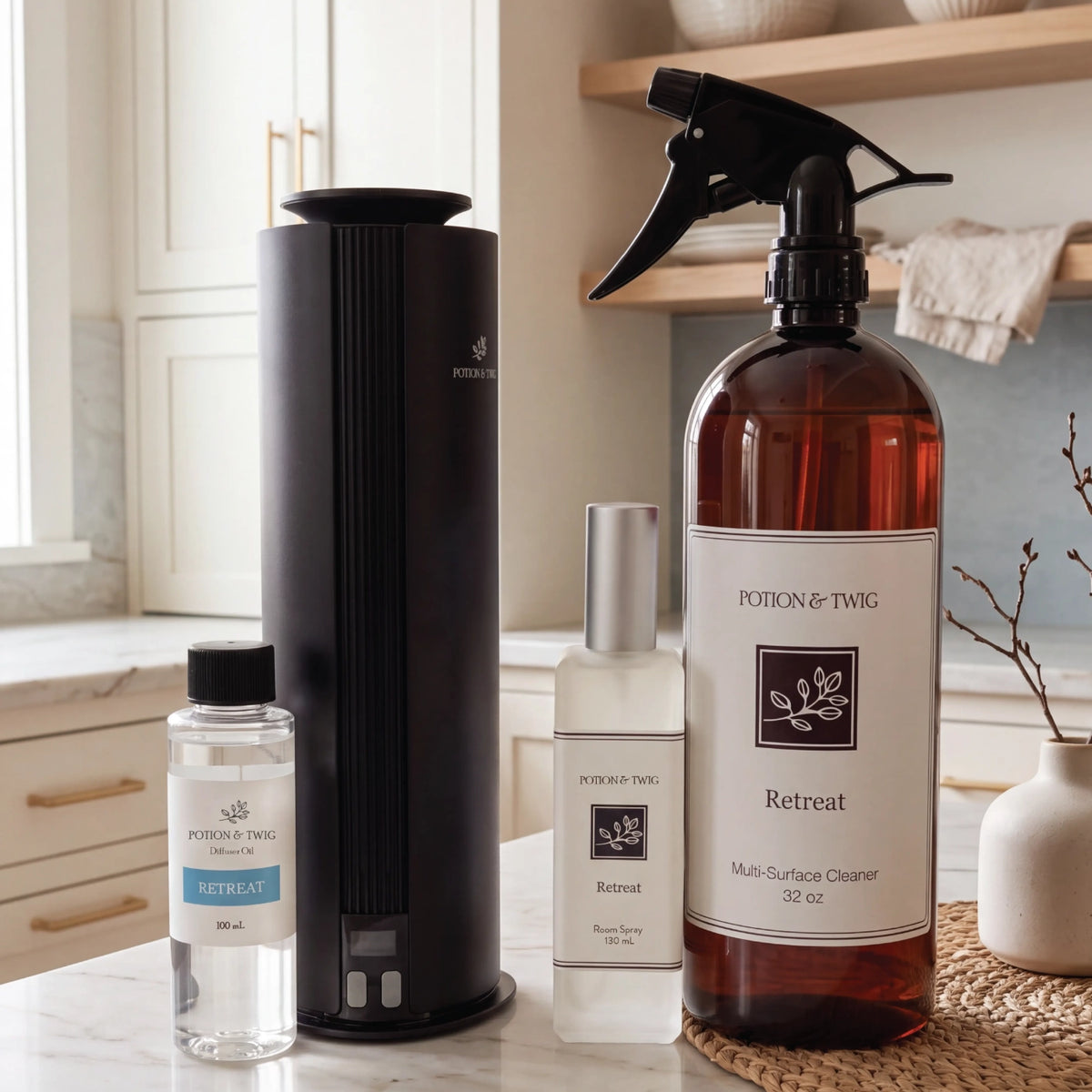 A set of Potion & Twig home fragrance products arranged on a marble countertop in a bright kitchen setting: a tall matte black diffuser, a small clear bottle labeled Retreat, a frosted bottle labeled Retreat, and a large amber spray bottle labeled Multi-Surface Cleaner.