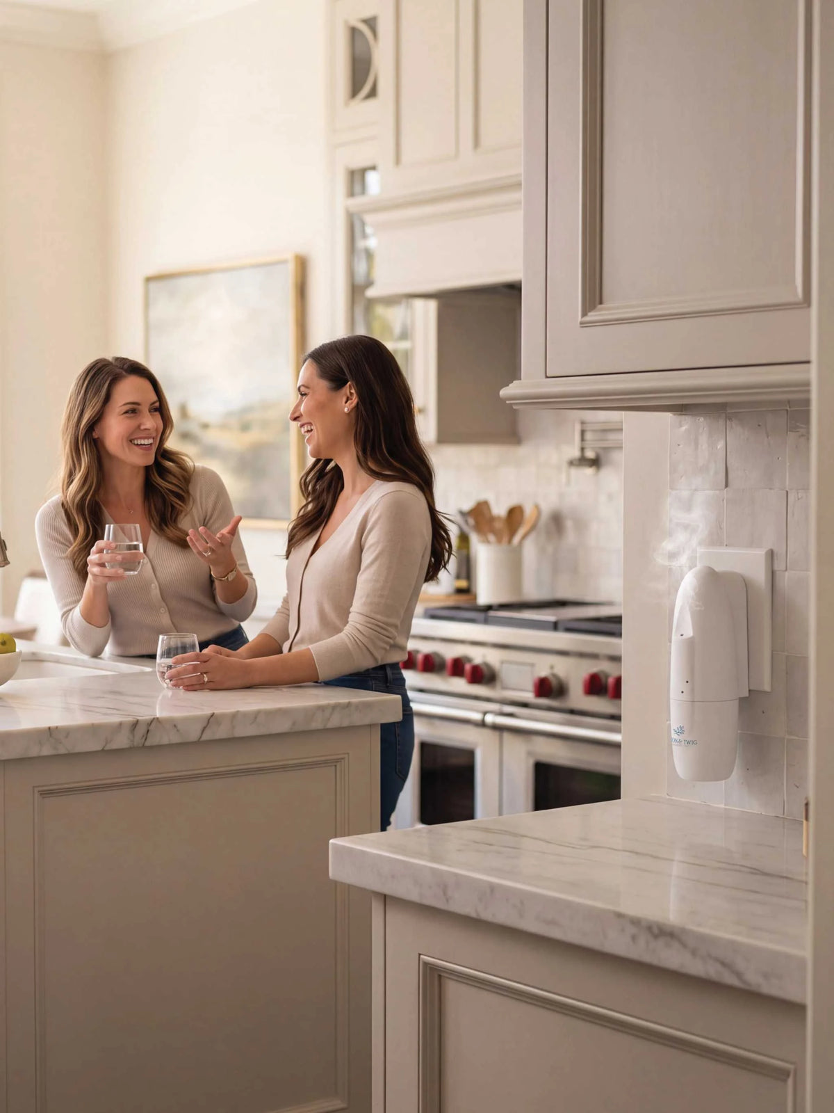 Two women smiling and conversing in a bright, modern kitchen with cream cabinetry and marble countertops. A white wall plug-in diffuser is visible on the right side of the kitchen wall.