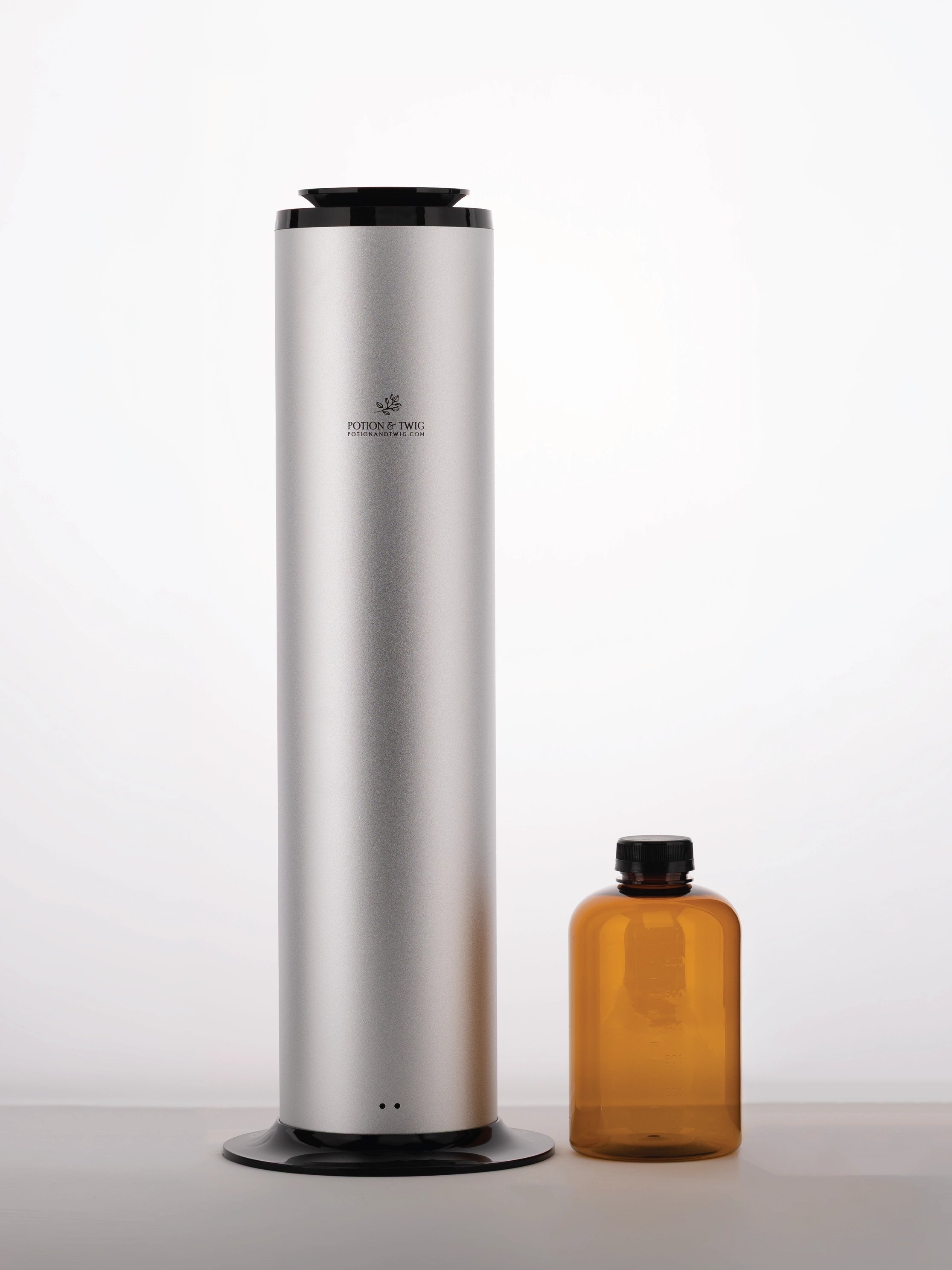 Tall silver cylindrical scent diffuser with an empty bottle of diffuser oil beside it on a white backrground