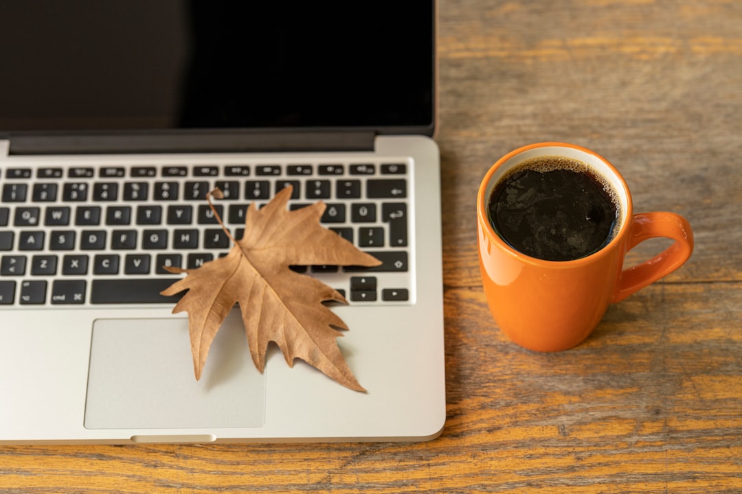 Silver laptop with a fall leaf on it. Sitting on a table with a fresh cup of coffee beside it.