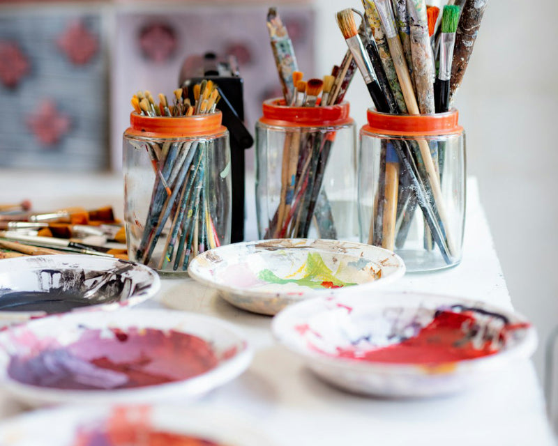 Three jars filled with paintbrushes on a table with art supplies.