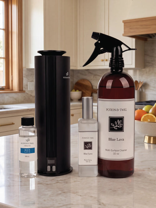 Potion & Twig home fragrance products displayed on a marble kitchen countertop, including a black cylindrical diffuser, a small clear bottle of fragrance oil, a frosted bottle with a silver cap, and a large amber spray bottle labeled Blue Lava.