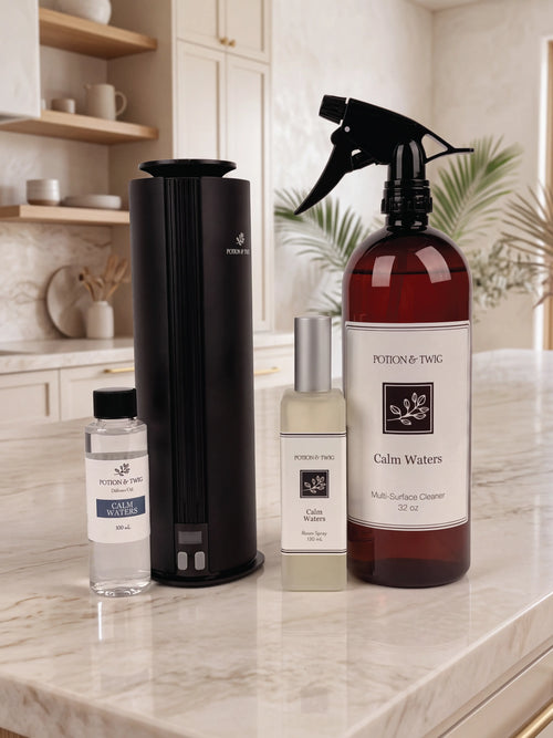 A set of Potion & Twig home care products arranged on a marble kitchen countertop, including a black cylindrical diffuser, a large amber spray bottle labeled Calm Waters Multi-Surface Cleaner, room spray bottle, and oil fragrance bottle