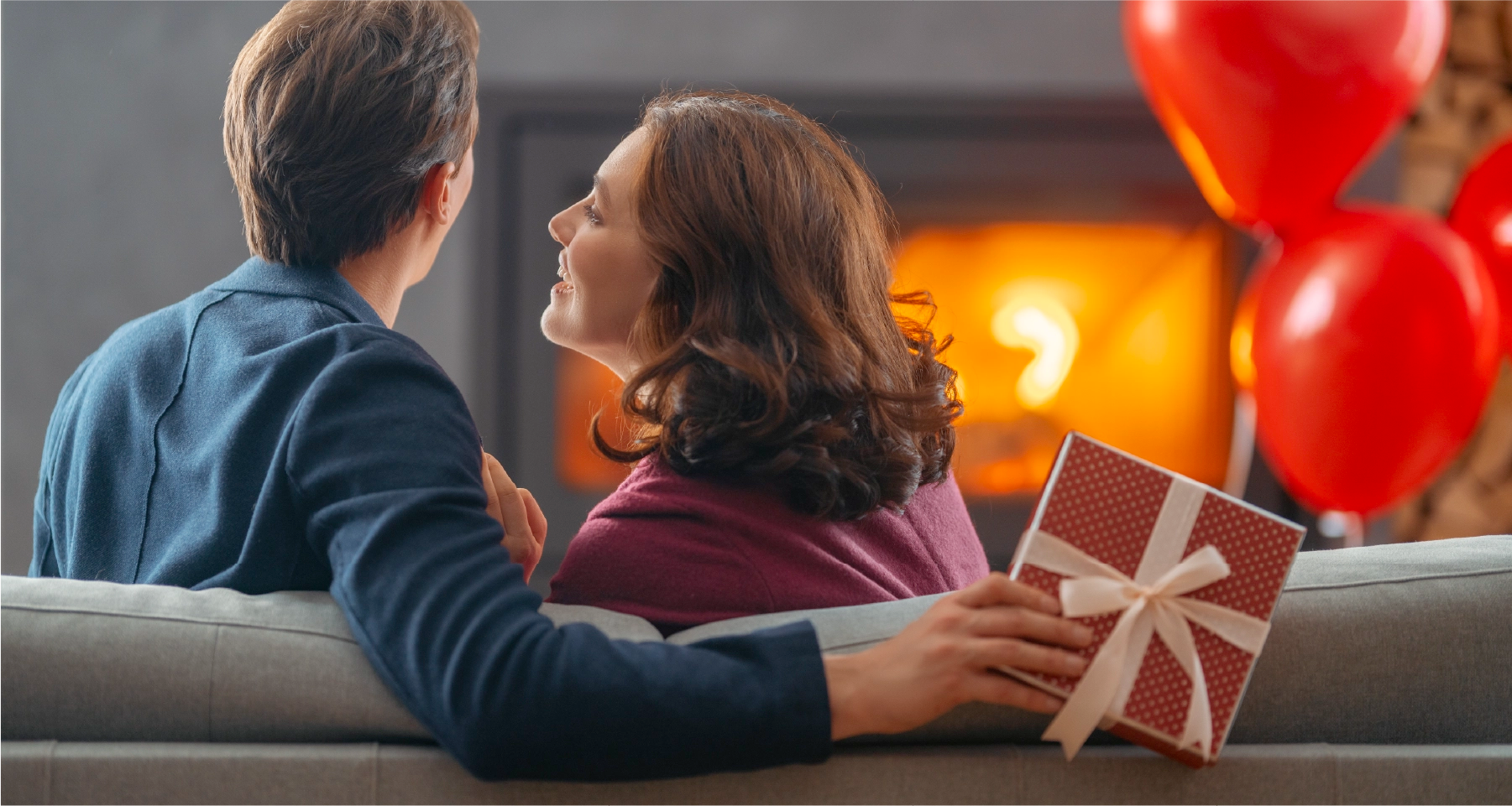Man and woman sitting on a couch with a gift and balloons, celebrating Valentine's Day.