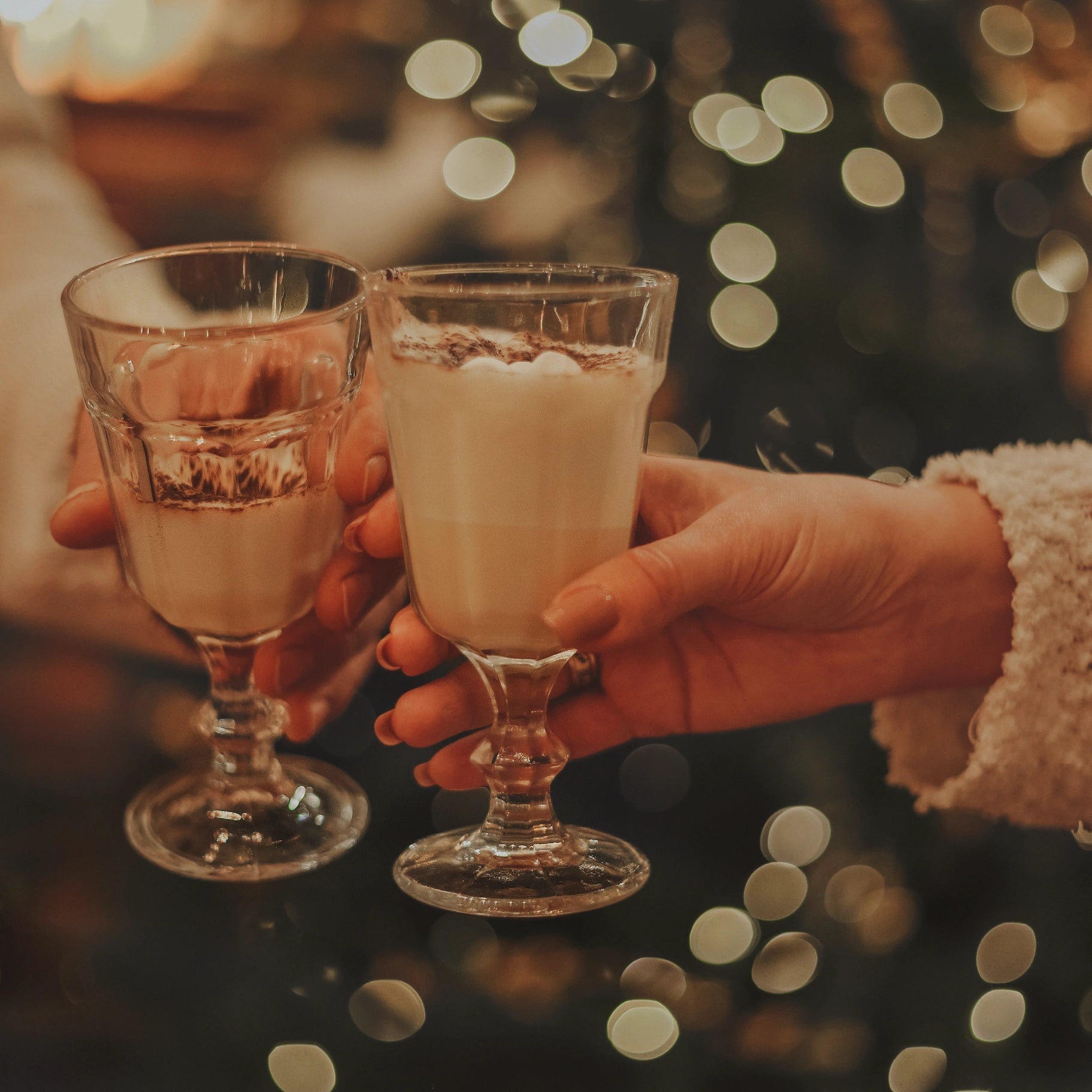 Two hands holding glasses of a creamy beverage against a blurred festive background with lights.