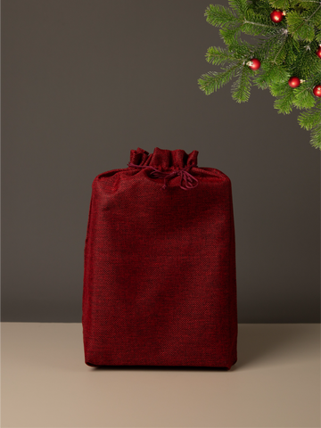 Red drawstring gift bag on a neutral background with a Christmas tree in the corner.