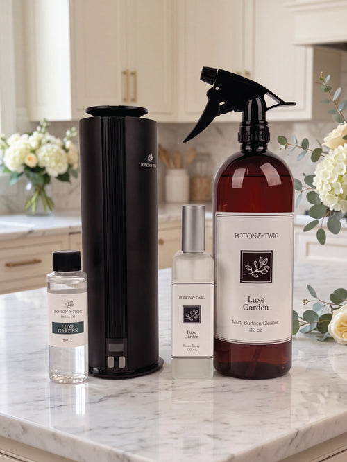 A home fragrance set from Potion & Twig arranged on a white marble countertop in a bright kitchen. The set includes a black cylindrical diffuser, a large amber spray bottle labeled Luxe Garden Multi-Surface Cleaner, a clear bottle of diffuser oil, and a clear spray bottle of Luxe Garden room spray.