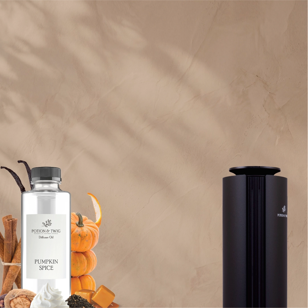A bottle of Pumpkin Spice diffuser oil by Poudrerie Doug next to a black cylindrical diffuser, styled with autumn spices and pumpkins against a textured beige wall with leaf shadows.