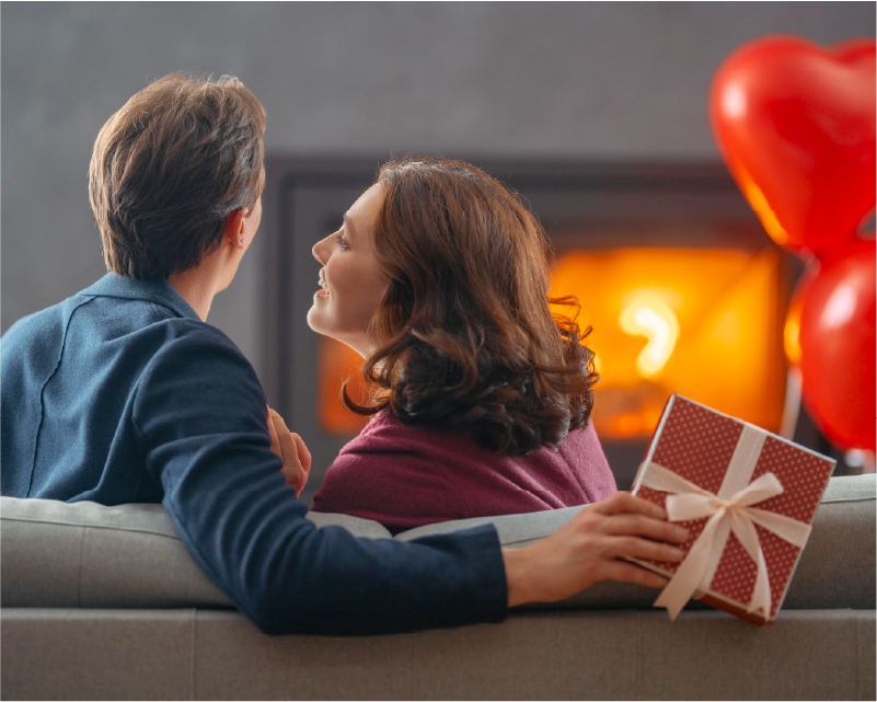 Man and woman sitting on a couch with a gift and balloons, celebrating Valentine's Day.