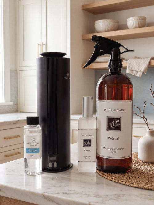 A home cleaning product set from Potion & Twig arranged on a marble kitchen counter, featuring a black cylindrical diffuser, a large amber spray bottle of Multi-Surface Cleaner, a small clear bottle of fragrance oil, and a small white bottle of a room spray.