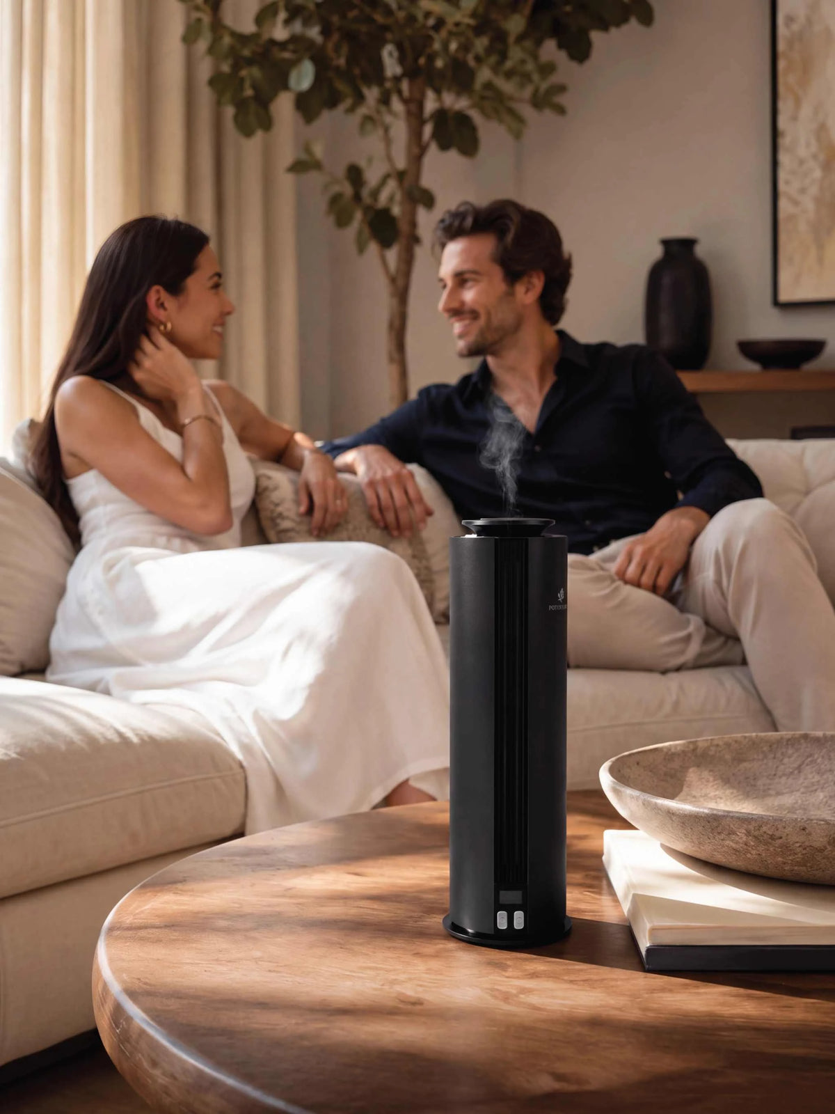 A black cylindrical tabletop diffuser with visible mist rising from the top, placed on a round wooden coffee table in a cozy living room. A man and a woman are seated on a cream sofa in the background, creating a warm, lifestyle-oriented scene.